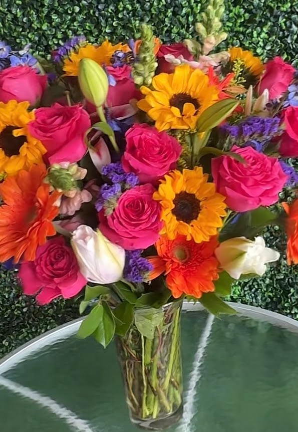 Colorful bouquet of pink roses, yellow sunflowers, white tulips, purple statice, and red gerbera daisies in a clear glass vase with green foliage