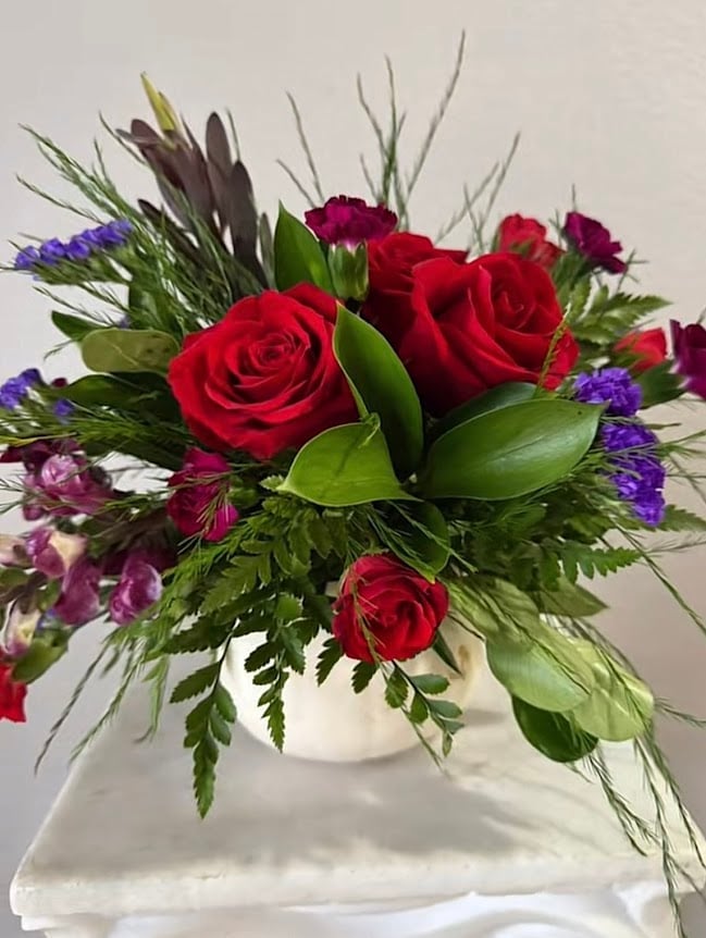 Vibrant floral arrangement with red roses, purple flowers, and green foliage in a white ceramic vase on white cloth
