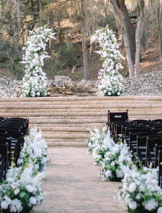 Wedding ceremony setup with white floral arrangements lining the aisle, black chairs, and a stone archway decorated with cascading white flowers against a wooded background