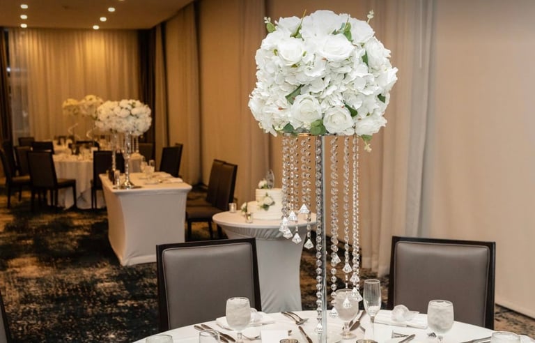 Elegant wedding reception venue with tall white hydrangea floral centerpiece on table, formal place settings, and draped crystal chandelier backdrop