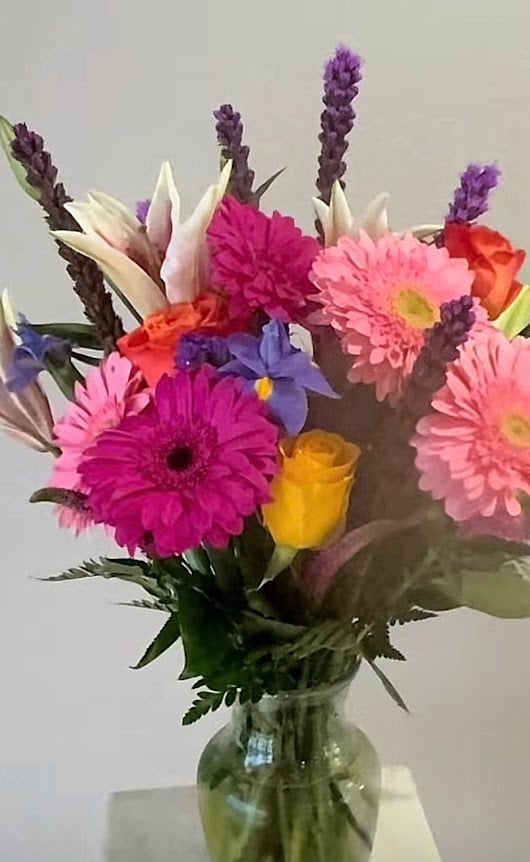Colorful flower arrangement in glass vase with pink and magenta gerberas, white lilies, yellow rose, and purple statice stems