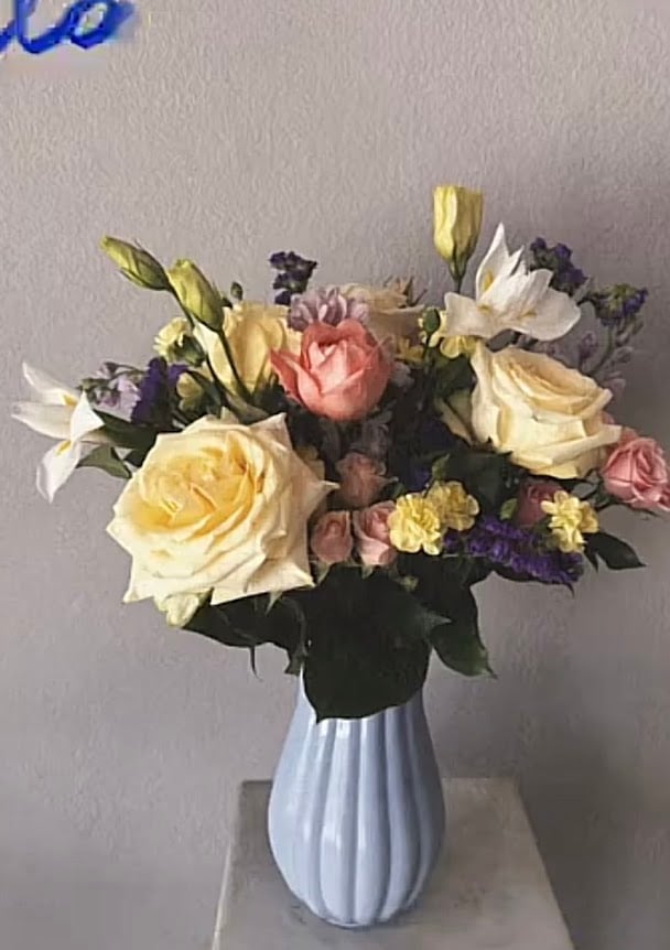 Mixed flower bouquet with yellow roses, coral peonies, white lilies, and purple accents in a light blue ribbed vase