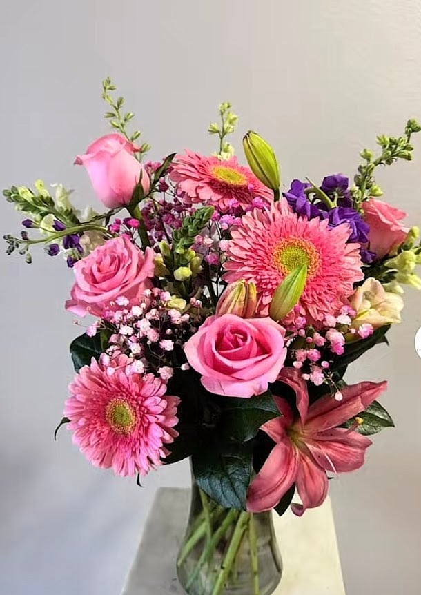 Vibrant flower bouquet in glass vase with pink roses, gerberas, purple flowers, and greenery on white background