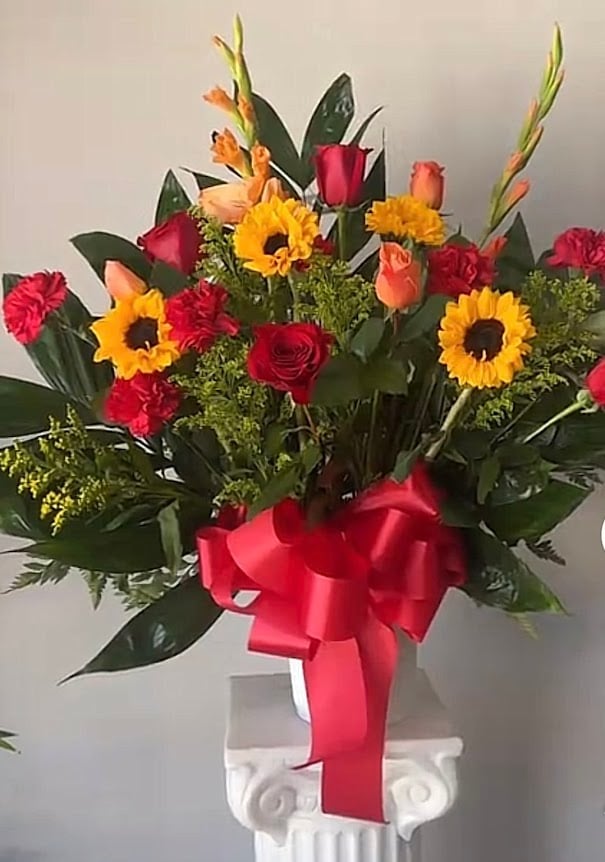 Colorful flower arrangement with yellow sunflowers, red roses and carnations, and orange tulips with green foliage in a white vase tied with a red ribbon bow